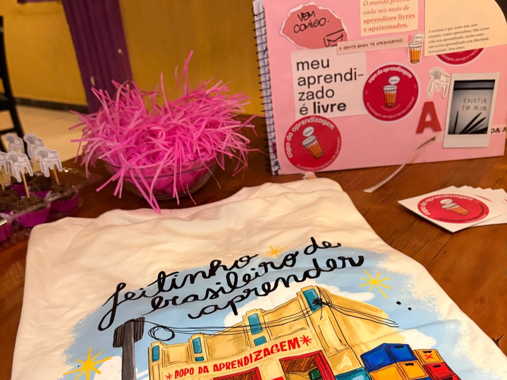 O caderno de colheitas do Copo aberto sobre a mesa, com colagens e a frase meu aprendizado é livre, ao lado de uma camiseta ilustrada do projeto.