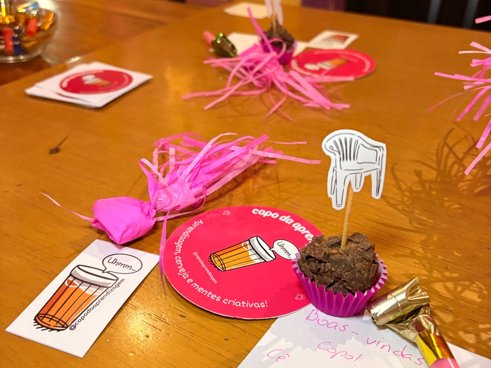 Detalhe da mesa: adesivo do Copo, bolacha de chope rosa, brigadeiro com uma cadeirinha de plástico espetada e um bilhete escrito à mão, boas-vindas copo, cê é fi de quem?
