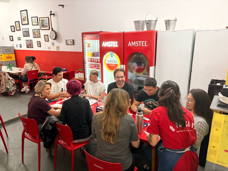 Grupo em volta de uma mesa vermelha de boteco, com geladeiras de cerveja ao fundo.