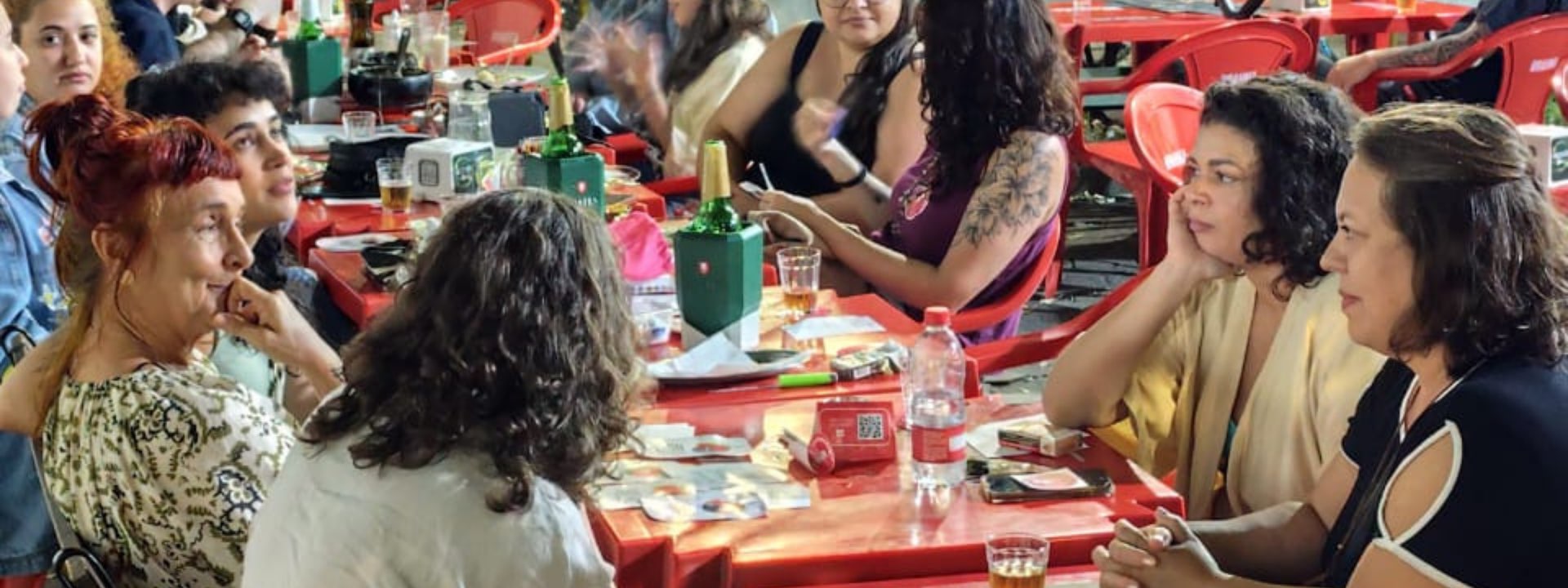 Mesa comprida de boteco cheia de gente conversando, com copos, cartinhas e artefatos espalhados sobre a mesa.
